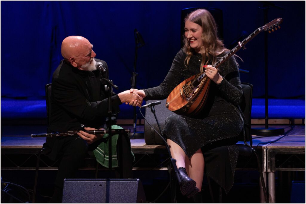 A man with a flute and a woman with a bouzouki shake hands and laugh on a stage with microphones in front of them.